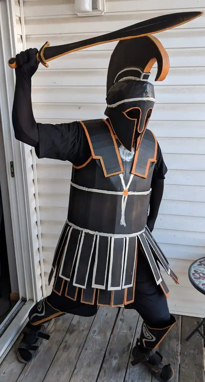 A person stands on a wooden porch in a homemade cardboard costume resembling a Spartan warrior, complete with helmet, chest…