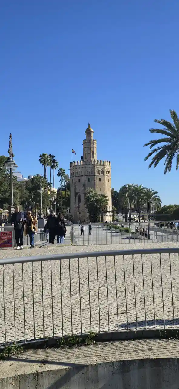A sunny photo of the historic Torre del Oro in Seville, Spain, with palm trees and pedestrians on a cobblestone walkway…