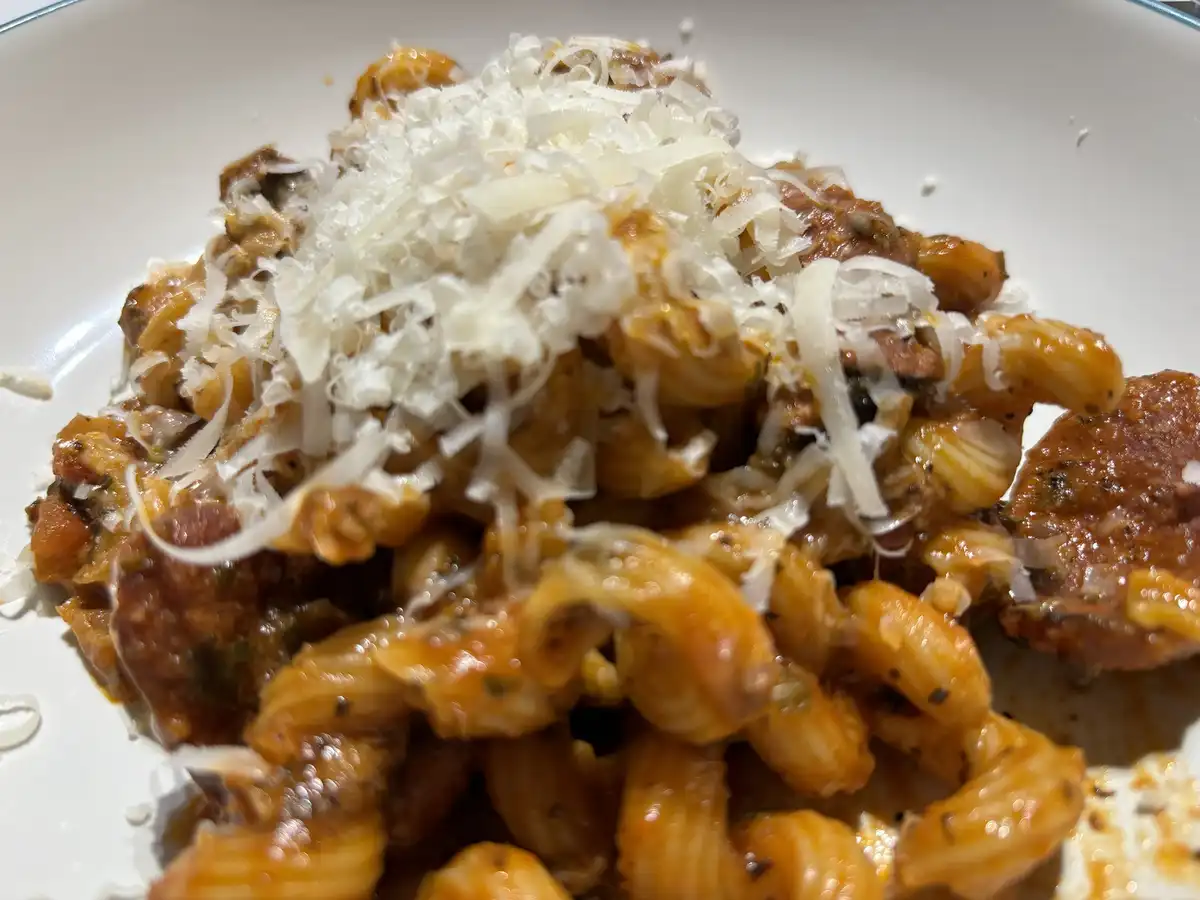 A close-up photo of a white plate piled high with pasta, likely rigatoni, coated in a rich red sauce and topped with a…
