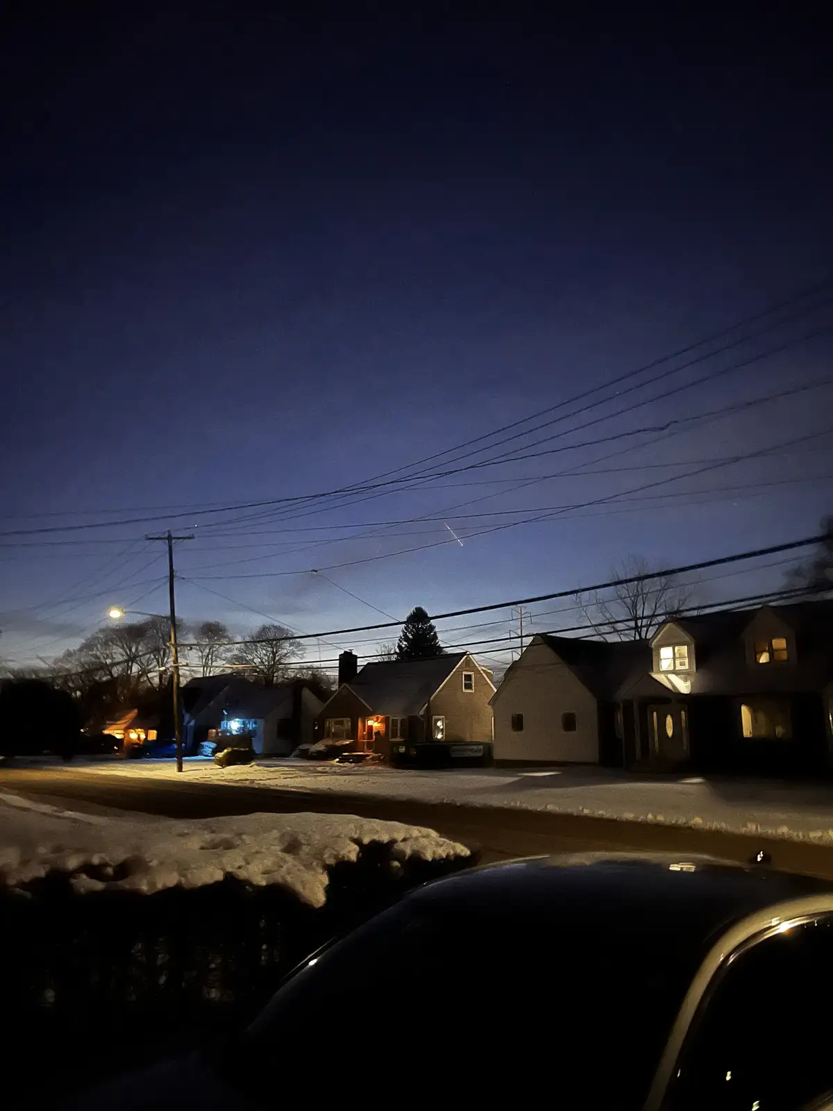 A nighttime photo of a snowy residential street with houses, power lines, and a car in the foreground. The sky is dark blue…