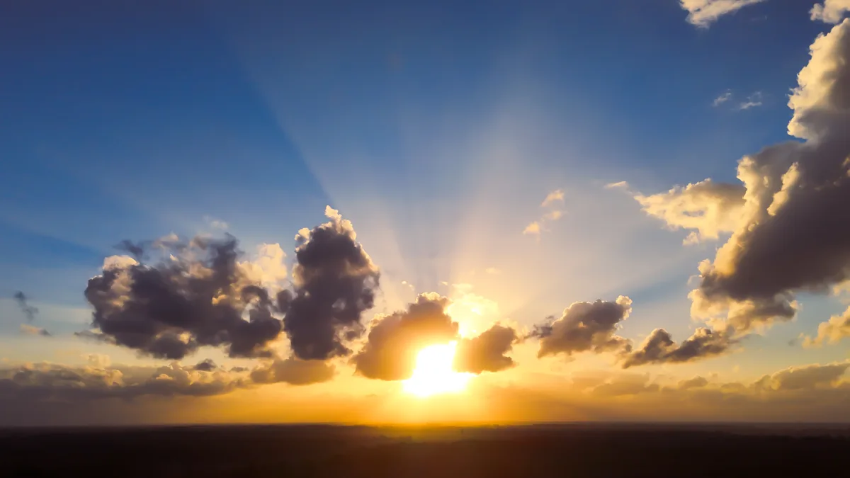 A dramatic sunset with the sun partially obscured by dark clouds, casting golden rays across a blue sky. The overlaid text…