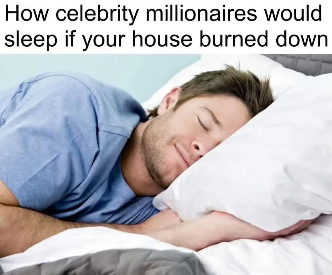 A man with short brown hair and a light beard is sleeping peacefully on his side in bed, wearing a blue t-shirt, with his…