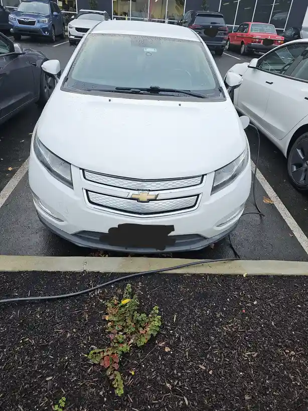 A white Chevrolet Volt is parked in a lot with a charging cable visibly unplugged from the car and lying on the ground. The…