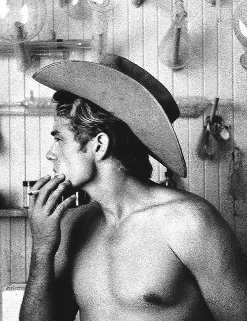 A black-and-white photo shows a shirtless man in profile wearing a cowboy hat and smoking a cigarette. He stands in a room…