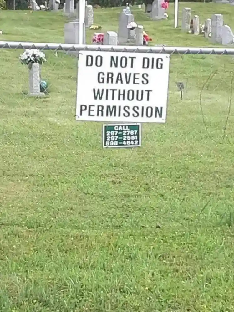 A white sign in a grassy cemetery warns 'DO NOT DIG GRAVES WITHOUT PERMISSION' and lists three phone numbers below it. The…