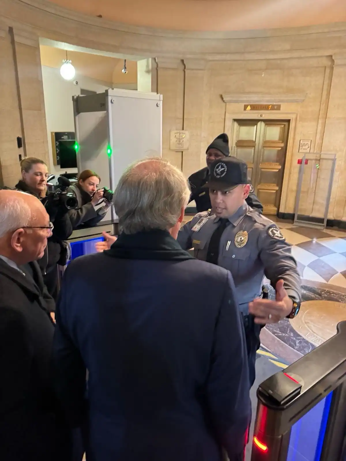 A security officer in a grey uniform and cap stands facing a man in a dark suit, gesturing firmly as if blocking his path…