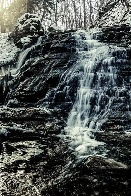 A long-exposure photograph of a snowy waterfall cascading over dark, layered rocks in a forest, with snow covering the…