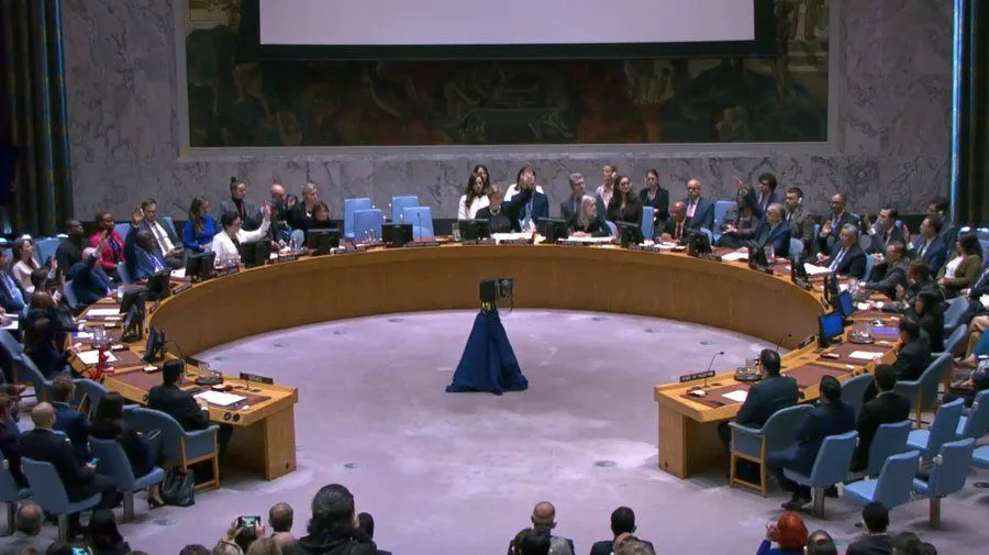 A wide shot of the United Nations Security Council chamber shows delegates seated around a large, curved wooden table. In…