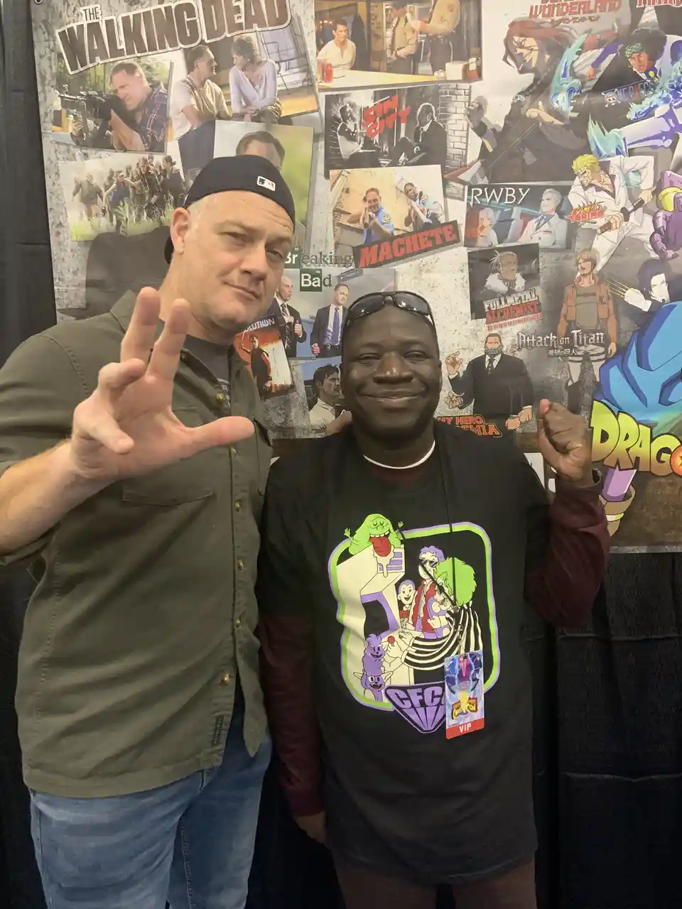 Two men pose in front of a wall covered with posters of popular TV shows and anime like The Walking Dead, Breaking Bad, Sin…