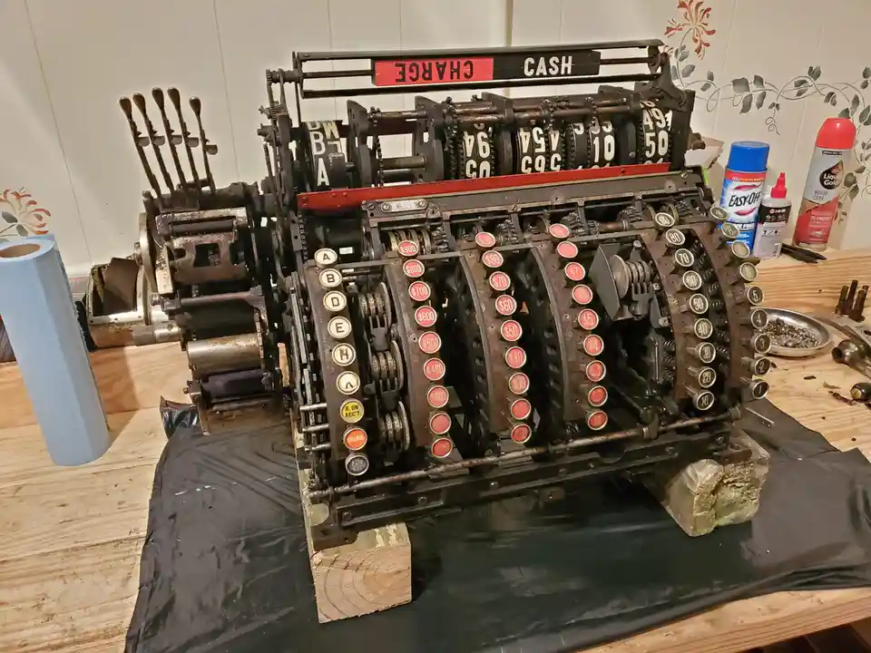 A heavily disassembled, rusted vintage mechanical cash register sits on a workbench, with its internal gears, number dials…