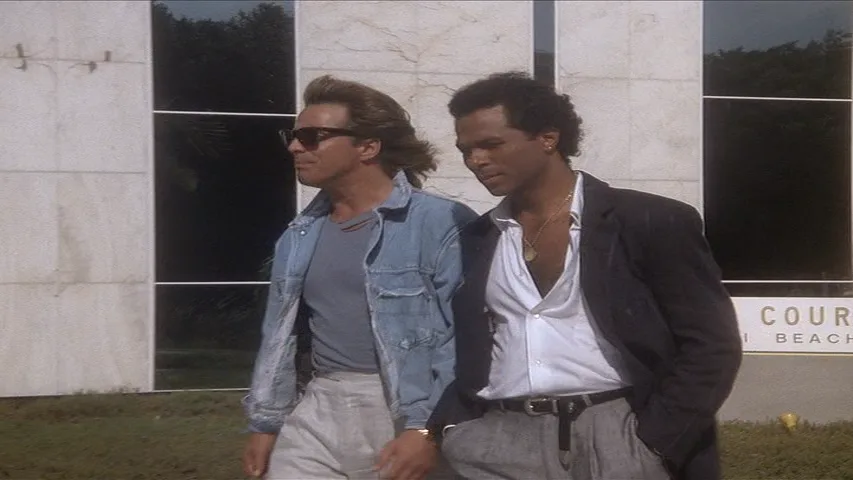 two men walk side by side outdoors, one in a denim jacket and sunglasses, the other in a dark blazer and white shirt, with a…