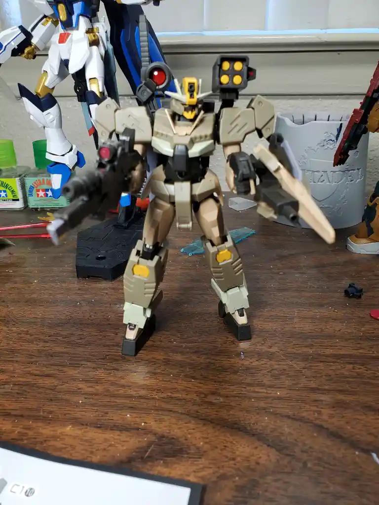 A tan and beige plastic robot model, resembling a Gundam, stands on a wooden desk surrounded by modeling supplies like glue…