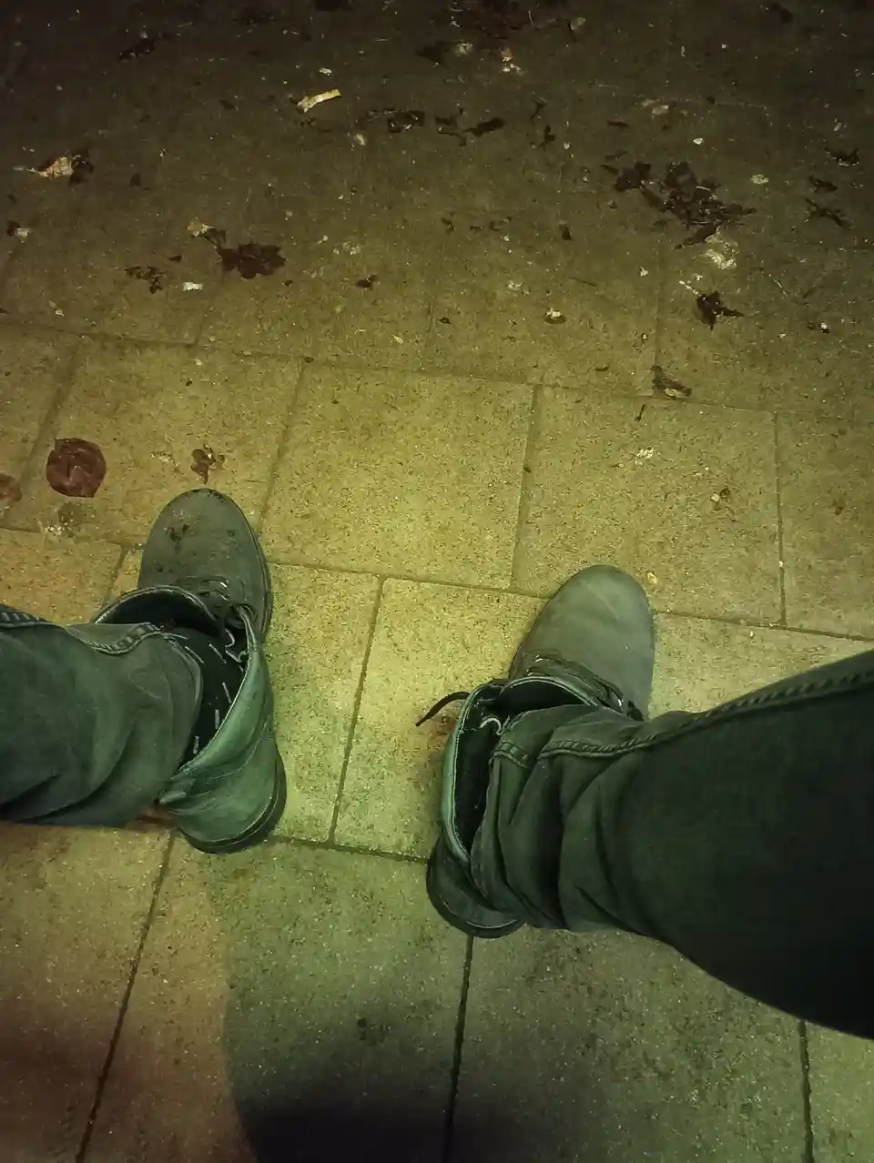 A first-person view of someone standing on a paved sidewalk at night, wearing dark green boots and dark jeans. The ground is…