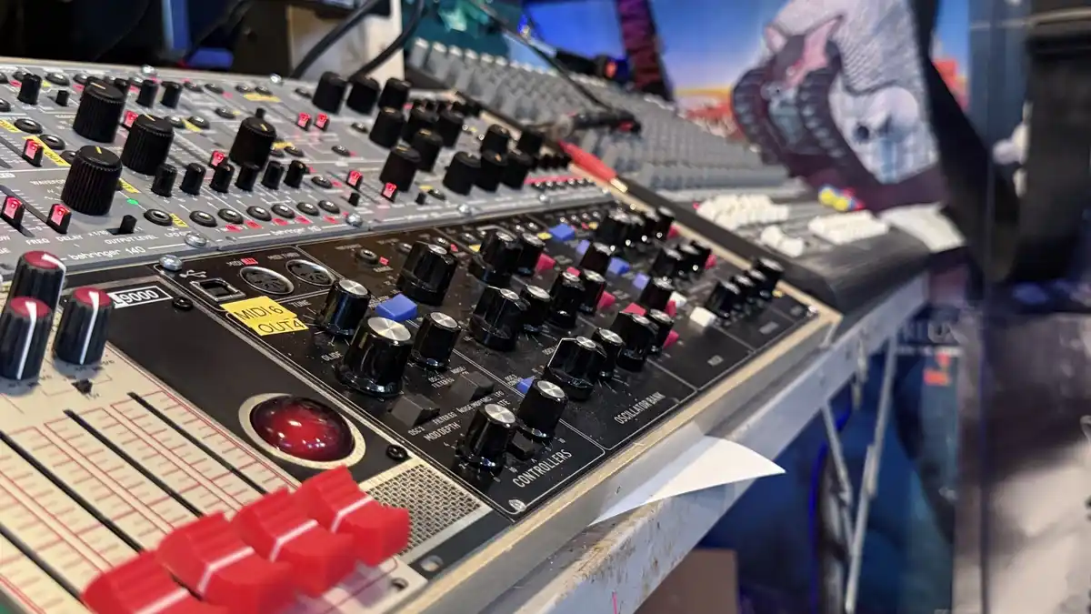 A close-up photo of a cluttered audio production desk with multiple synthesizers and mixers, including a Behringer mixer and…