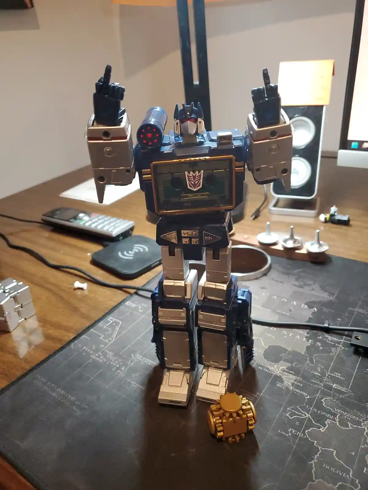 A blue and silver Transformers toy robot stands on a desk with a world map mousepad, its arms raised with fingers pointing…