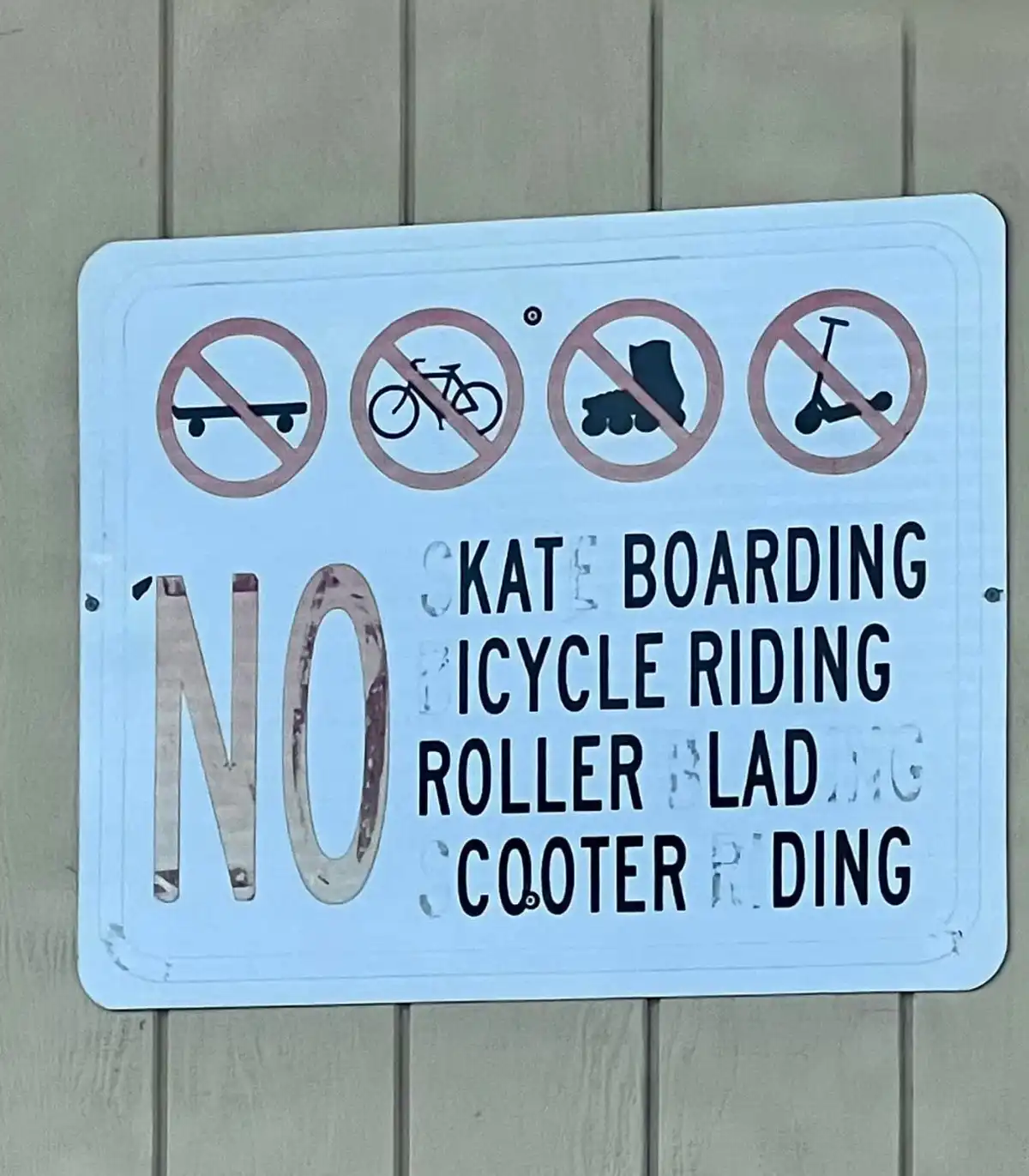A weathered white sign with four red prohibition symbols above text that reads 'NO KATE BOARDING BICYCLE RIDING ROLLER BLAD…