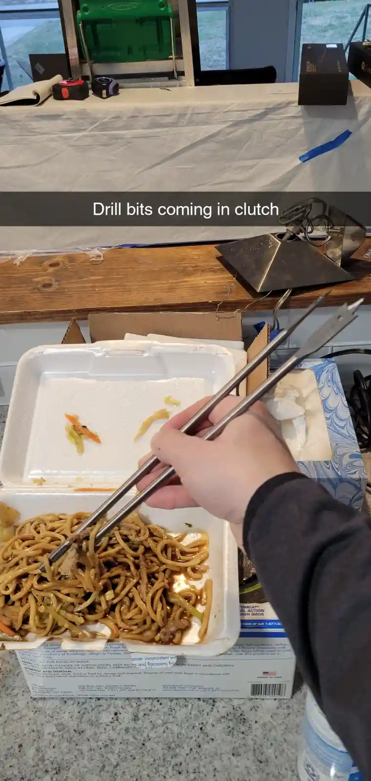 A hand holds metal chopsticks, which are actually drill bits, over a nearly empty takeout container of noodles. The text…