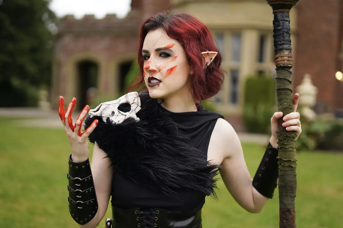 A woman with red hair, pointed elf ears, and dark makeup holds a staff and a skull, with red paint on her face and fingers…
