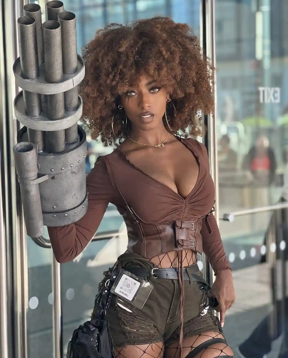 A woman with voluminous curly hair poses confidently while holding a large, multi-barreled machine gun prop. She wears a…