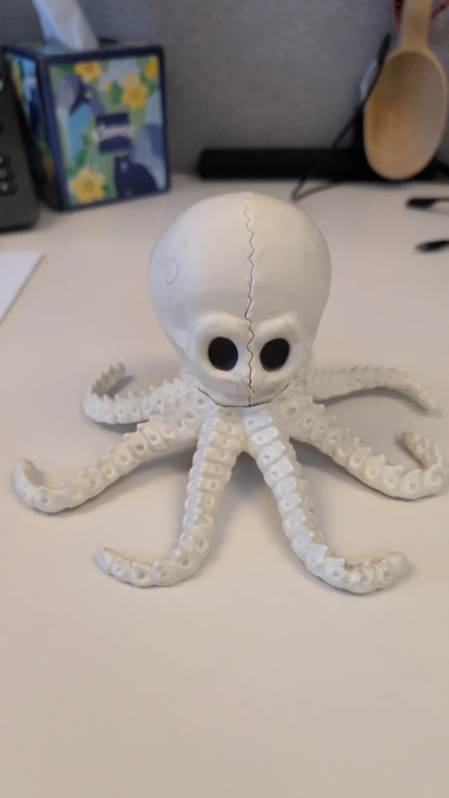 A white plastic toy octopus sits on a desk, its head sculpted to resemble a human skull with large black eye sockets and a…