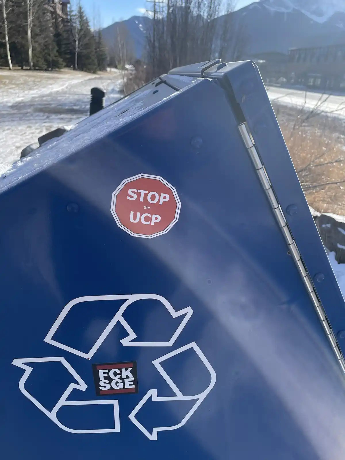 A close-up of a blue recycling bin with snow on its lid, featuring a red stop sign sticker that reads 'STOP the UCP' and a…