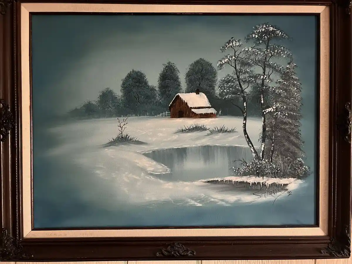 A framed painting depicts a serene winter scene with a snow-covered wooden cabin, frozen pond, and snow-laden trees under a…