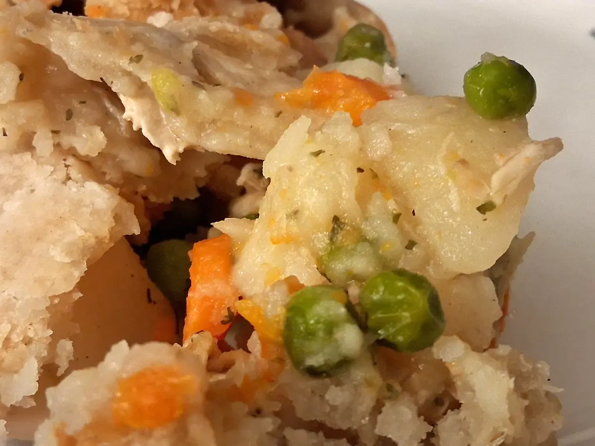A close-up photo of a savory dish with mashed potatoes, peas, and carrots, showing a textured, home-cooked appearance. The…