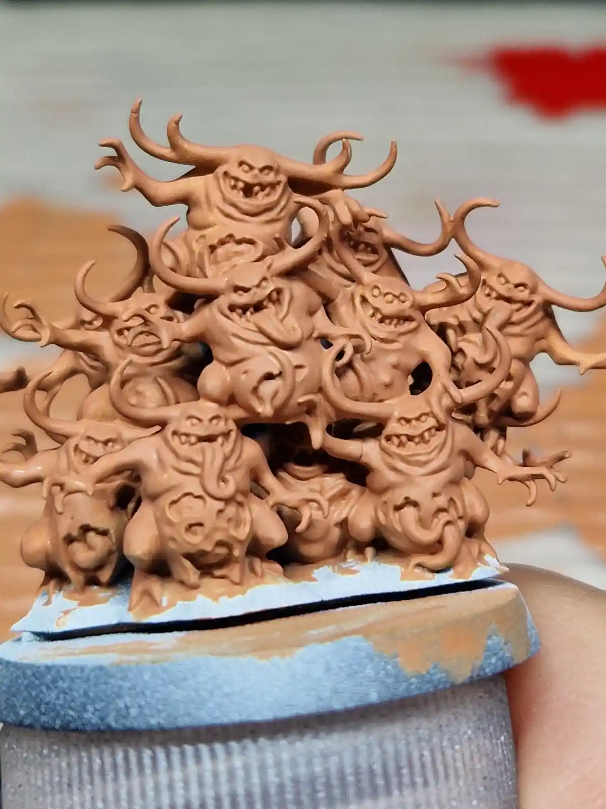 A close-up photo shows a hand holding a miniature wargaming model of a chaotic pile of grotesque, horned, grinning monsters…