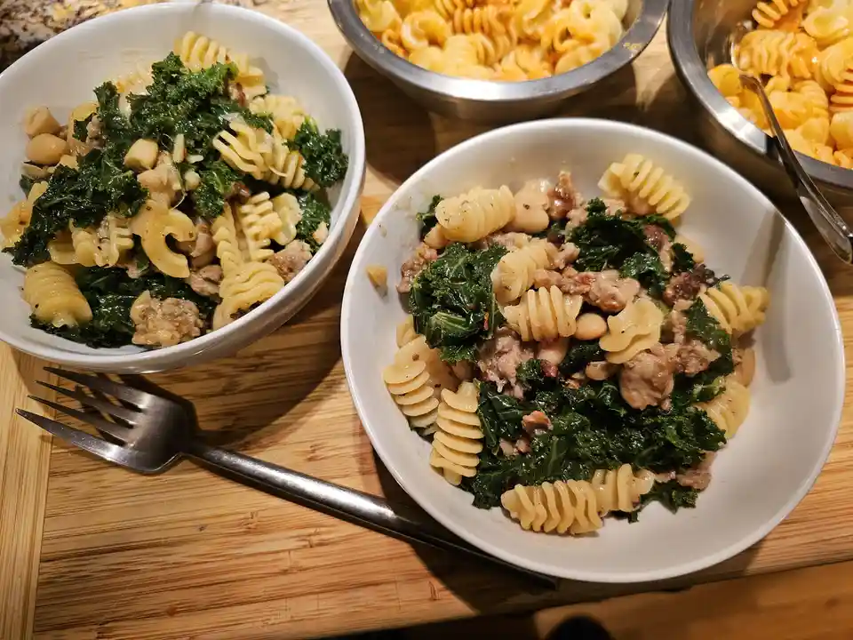 Two white bowls filled with spiral pasta, kale, white beans, and crumbled sausage sit on a wooden surface, with a metal fork…