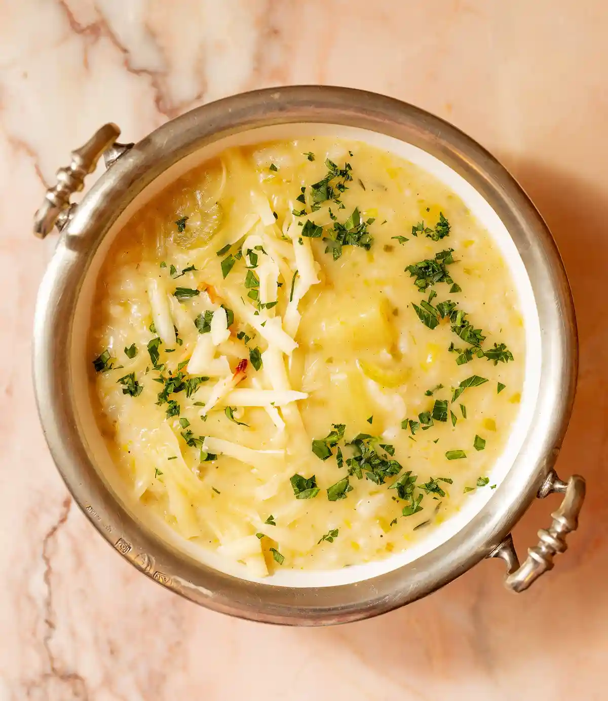 A top-down photo of a creamy, yellowish soup or risotto in a silver bowl with ornate handles, garnished with shredded white…