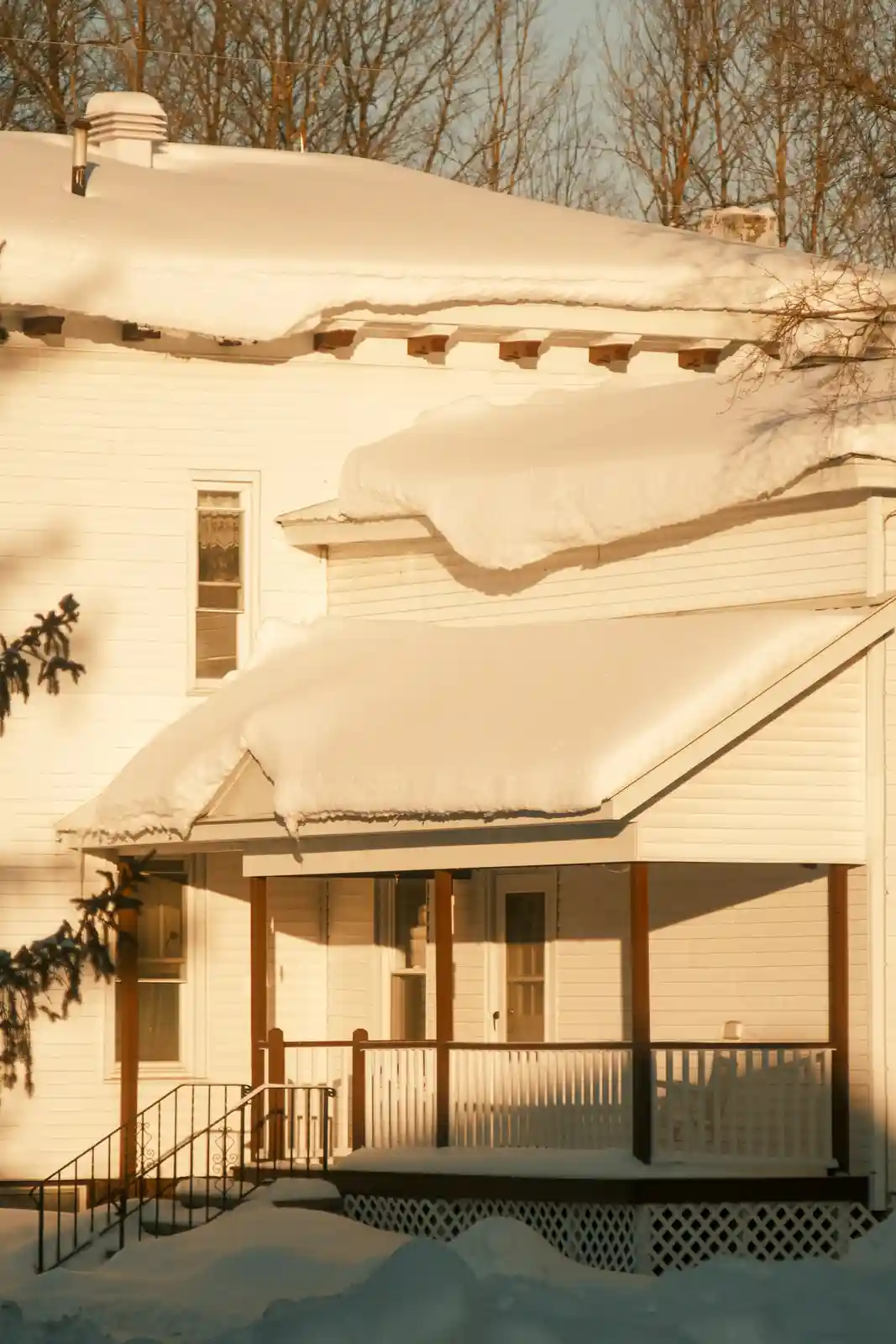 A white house covered in thick snow, with massive snowdrifts piled high on the roof and porch, illuminated by warm sunlight…
