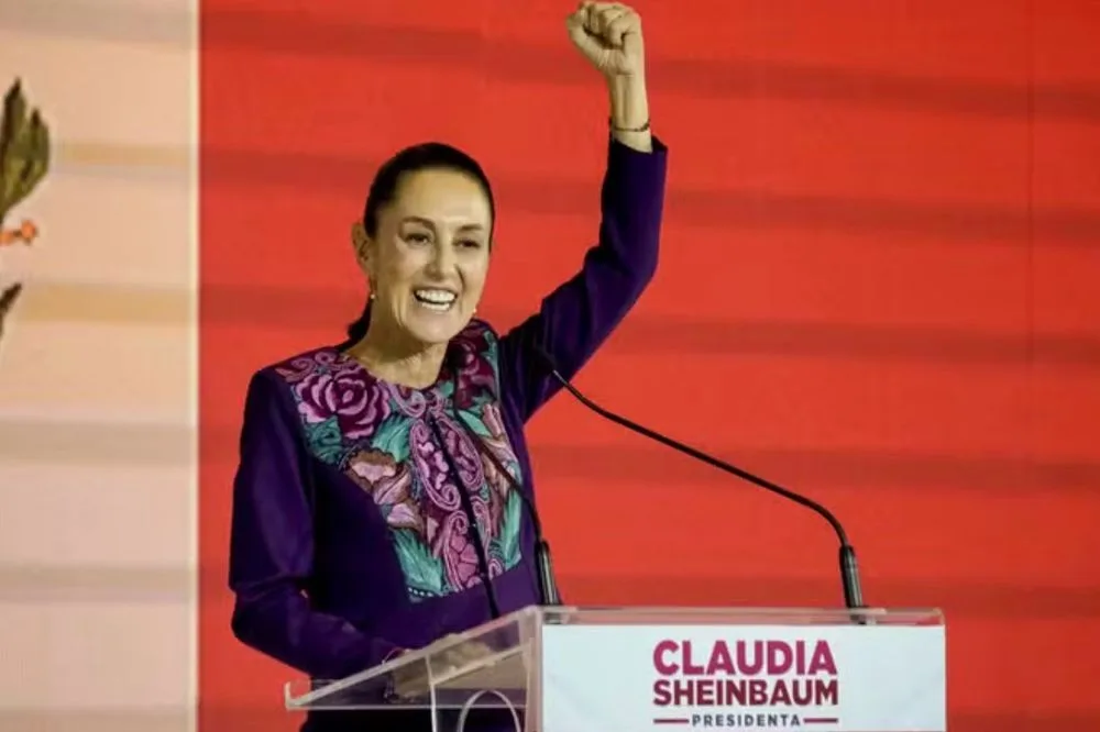 A woman stands at a podium with a sign reading 'CLAUDIA SHEINBAUM PRESIDENTA', raising her right fist in a triumphant…