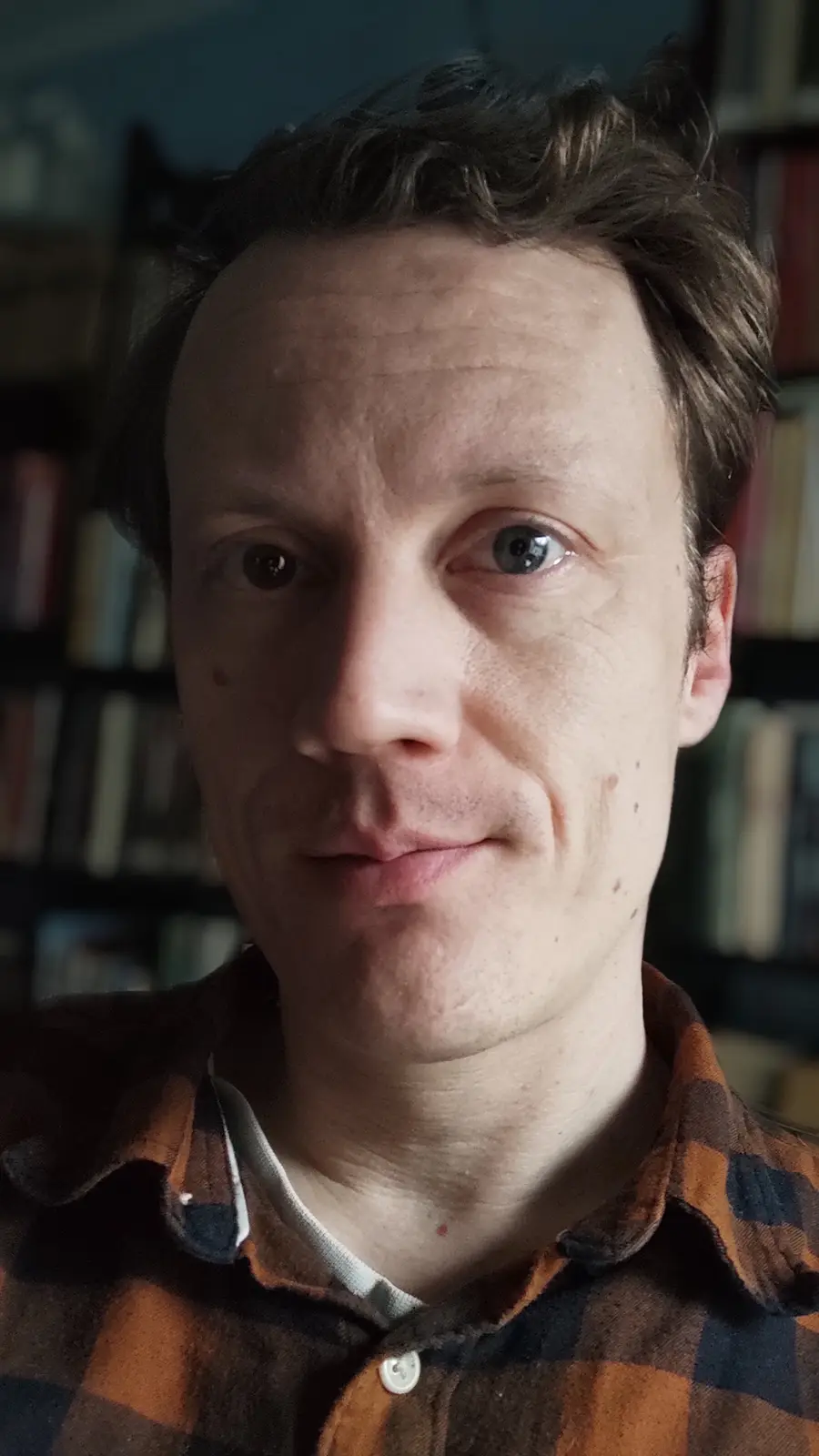 A close-up photo of a man with dark hair and blue eyes, wearing a brown and orange plaid shirt, looking directly at the…