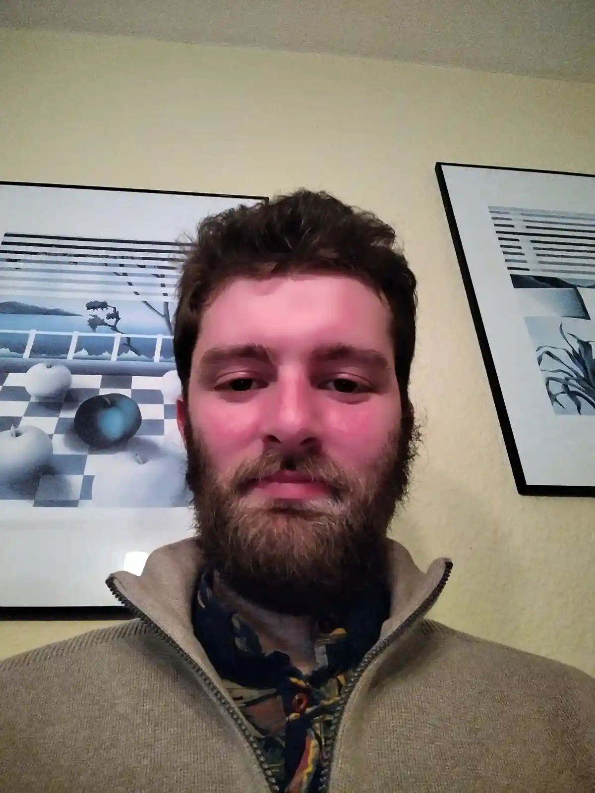 A man with a beard and messy hair stares directly at the camera with a neutral expression, wearing a beige zip-up sweater…