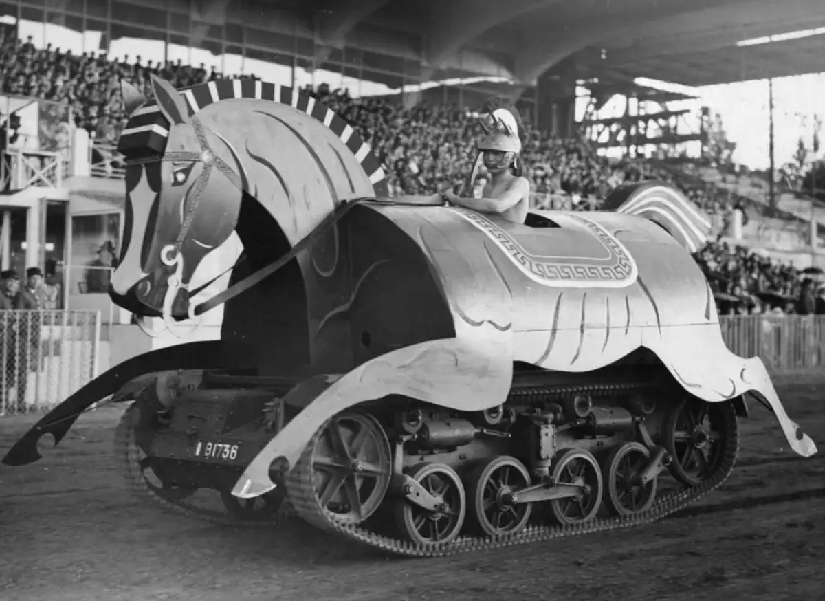 A black-and-white photo shows a large, ornate wooden horse with tank treads and a driver in a helmet, set in a stadium with…
