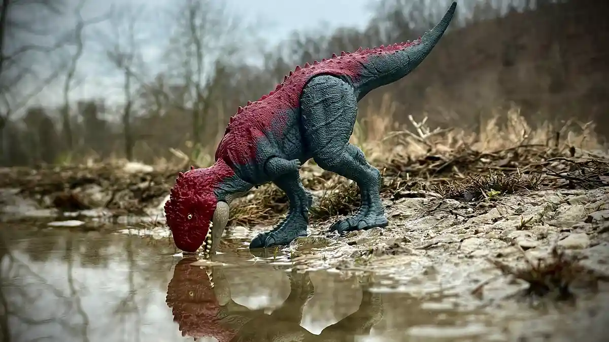 A plastic toy dinosaur with a red head and back and grey body is positioned at the edge of a shallow puddle in a natural…