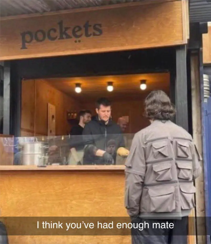 A person wearing a jacket with many pockets stands at a food stall named 'pockets'. The staff member behind the counter…