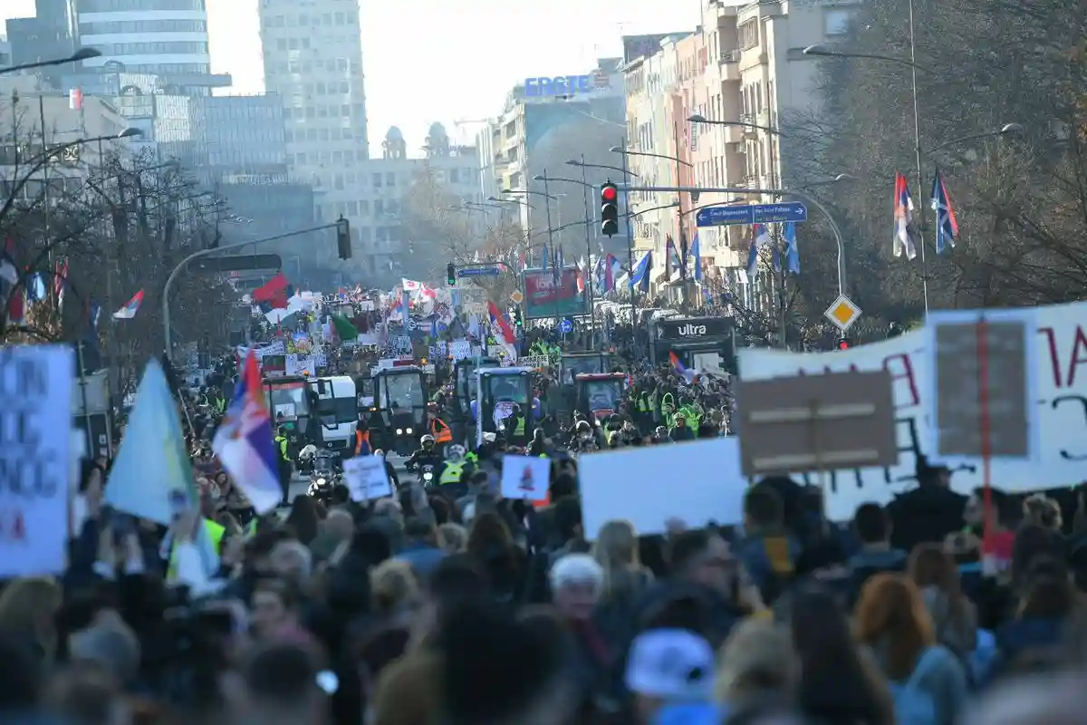 A large protest march fills a city street, with thousands of people holding signs and waving flags, including Serbian flags…