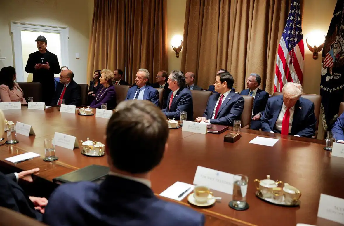 A photo of a formal meeting with several officials seated around a large wooden table, including Donald Trump seated at the…