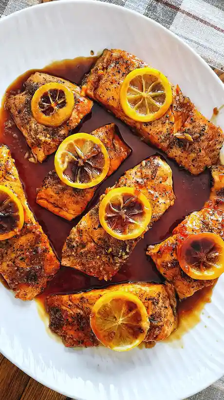 A white plate holds several cooked salmon fillets glazed in a dark sauce and topped with caramelized lemon slices. The text…