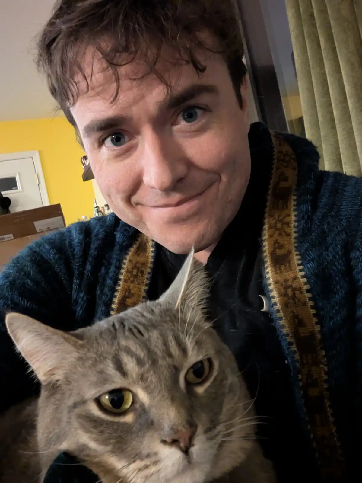 A man with blue eyes and a slight smirk holds a gray tabby cat close to the camera. The cat has wide, unimpressed yellow…