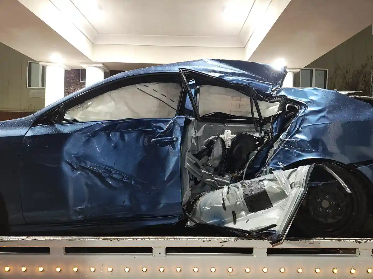 A severely damaged blue sedan, with its front end crushed and passenger side door open, sits on the flatbed of a tow truck…