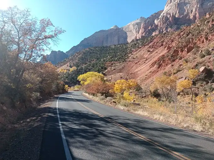 A scenic road winds through a canyon with red rock formations and autumn-colored trees under a clear blue sky. Overlaid text…