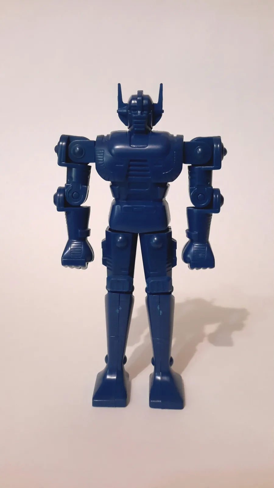A solid blue plastic robot toy stands upright against a plain off-white background, facing forward with arms at its sides…