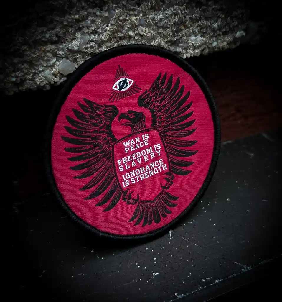 A circular red patch with a black embroidered double-headed eagle holding a shield. Above the eagle is an all-seeing eye…