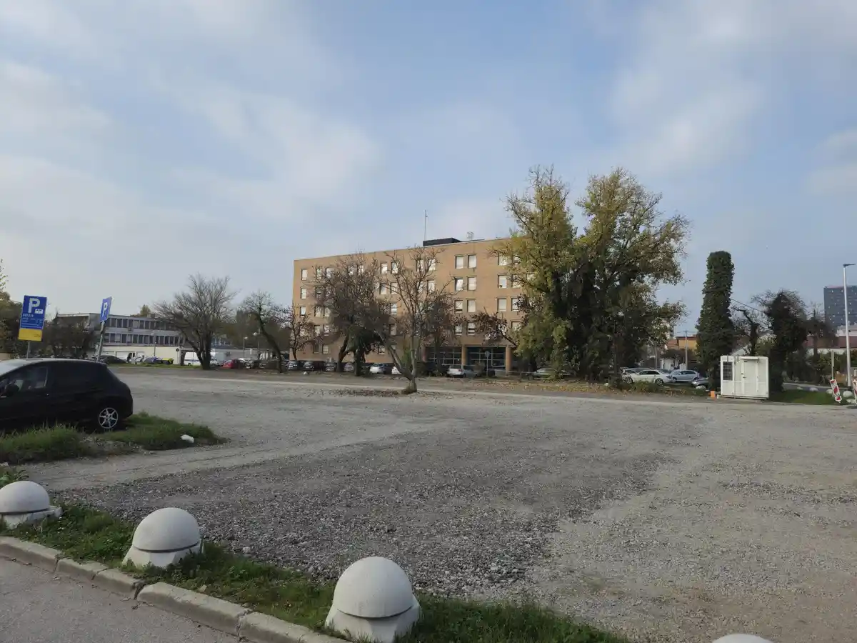 A gravel parking lot with a multi-story brick building in the background, surrounded by trees with autumn leaves. Several…