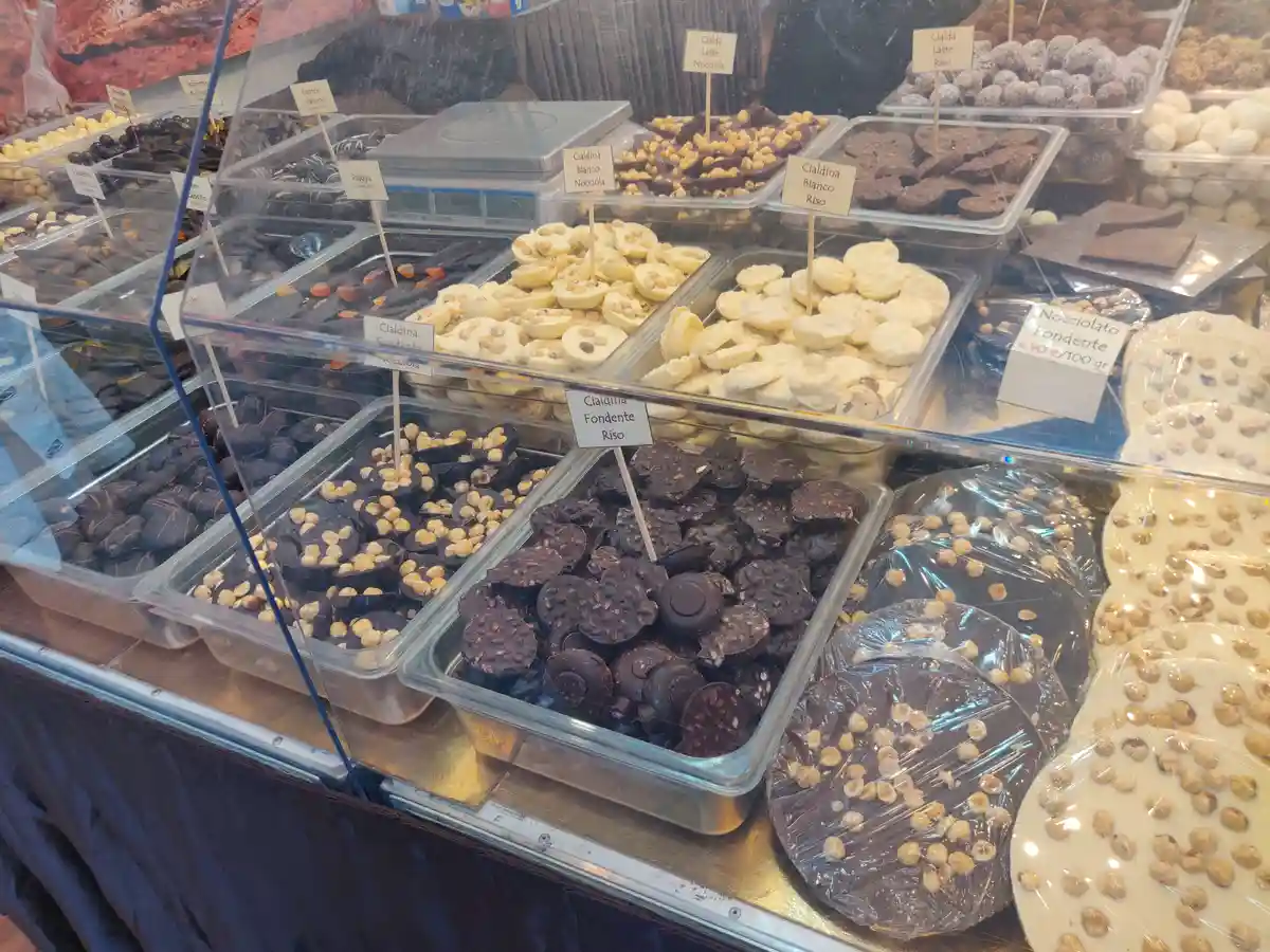 A display case filled with various types of chocolate confections in clear bins, each labeled with small signs indicating…