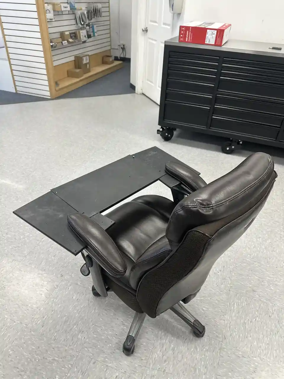 A brown leather ergonomic office chair with a large, flat, black metal desk surface bolted directly to its armrests and…