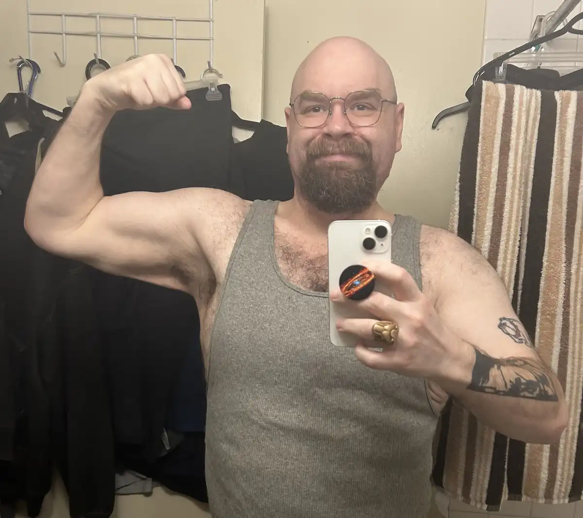 A bald man with glasses and a beard takes a mirror selfie in a bathroom, flexing his bicep while holding a white smartphone…