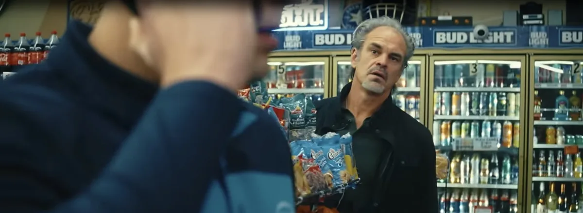 A man with gray hair and a black jacket stands in a convenience store, holding a shopping basket full of snacks, looking…